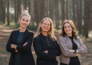 Der Vorstand des Kunstvereins Haan: Drei Frauen stehen lächelnd mit verschränkten Armen nebeneinander in einem sonnendurchfluteten Wald. Patrycja Karle, Pia Stojkovic und Frauke Wagener präsentieren sich als neues Leitungsteam – selbstbewusst, offen und nahbar.