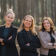 Der Vorstand des Kunstvereins Haan: Drei Frauen stehen lächelnd mit verschränkten Armen nebeneinander in einem sonnendurchfluteten Wald. Patrycja Karle, Pia Stojkovic und Frauke Wagener präsentieren sich als neues Leitungsteam – selbstbewusst, offen und nahbar.