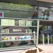 Ausstellung des Kunstvereins Haan im Schaufenster der Stadtbücherei Haan: Zu sehen sind abstrakte Gemälde, Fotografien und grafische Arbeiten in drei Reihen. Links oben das Logo des Kunstvereins.